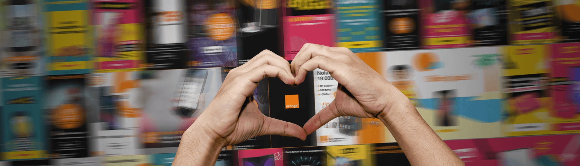 Deux mains forment un coeur devant un mur de publicités digitales pour l'opérateur Orange