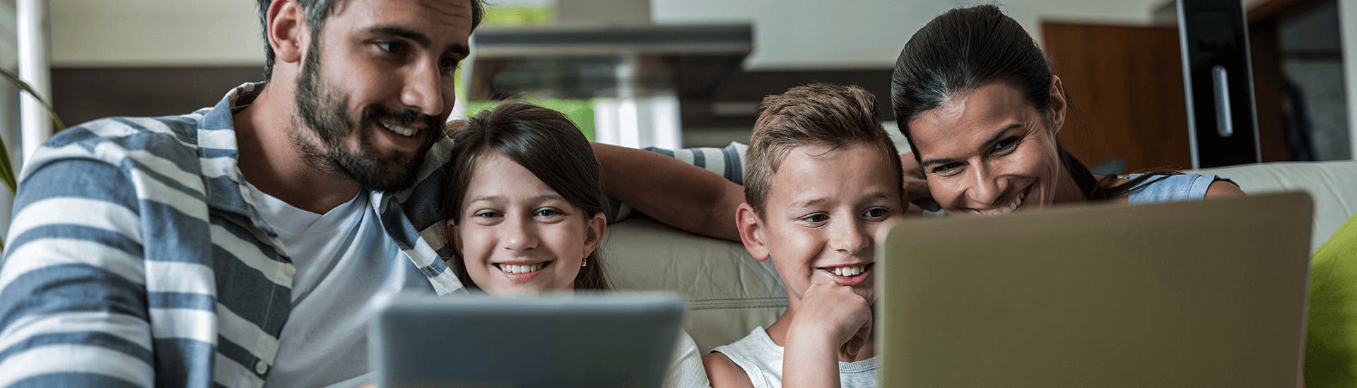 Une famille ensemble sur un canapé regarde en souriant un ordinateur portable et une tablette